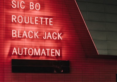Red neon letters are mounted on a dark tiled wall, displaying the words Poker, Sic Bo, Roulette, Black Jack, and Automaten.