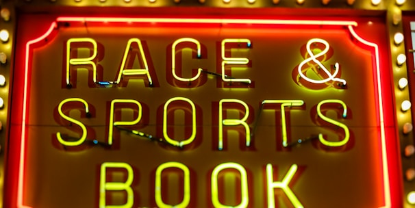 Bright neon lights on a marquee sign display the words 'RACE & SPORTS BOOK'. The sign is surrounded by rows of yellow and white lights against a red background, evoking a vibrant, energetic atmosphere typically associated with casinos or entertainment venues.