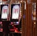 A pair of casino slot machines, each with detailed screens displaying various game outcomes and jackpot amounts. The chairs in front of the slot machines are dark and cushioned for comfort. The background features a hanging curtain made of golden ropes and other casino decorations are visible.