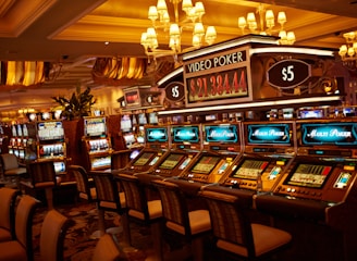 A casino floor featuring rows of brightly lit video poker machines. The area is illuminated by elegant chandeliers, with a display board showing a large sum of money, suggesting a possible jackpot. The rich decor includes luxurious carpets and comfortable seating, adding to the opulent atmosphere.
