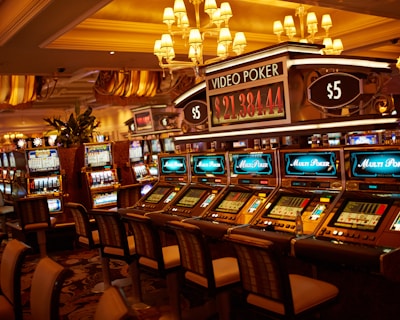 A casino floor featuring rows of brightly lit video poker machines. The area is illuminated by elegant chandeliers, with a display board showing a large sum of money, suggesting a possible jackpot. The rich decor includes luxurious carpets and comfortable seating, adding to the opulent atmosphere.