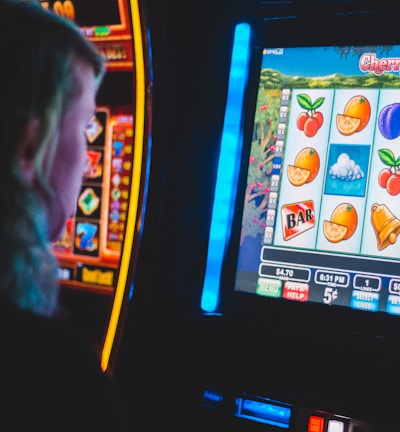 A person is closely observing a digital slot machine with colorful fruit symbols, bars, and a bell. The slot machine screen displays symbols like oranges, plums, cherries, and clouds. The background is dimly lit, creating a focused and somewhat intense atmosphere.