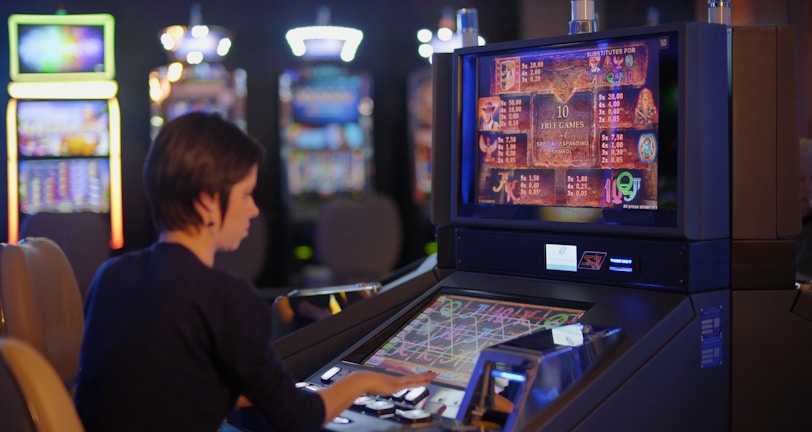 A person is seated at a slot machine console in a casino, focusing intently on the game displayed on the screen. The background shows several other illuminated gaming machines, adding a vibrant and colorful atmosphere to the scene.