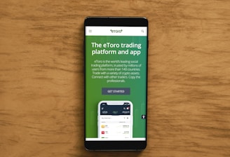 A smartphone displaying an advertisement for an online trading platform called eToro. The screen shows a green background with text promoting the platform as a leading social trading platform. A button labeled 'Get Started' is visible, along with a partial view of a mobile app screen below.