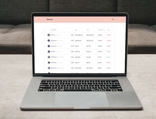 A laptop on a table displaying a cryptocurrency trading platform. The screen lists various cryptocurrencies along with their buy and sell values, spread percentages, and 24-hour change percentages. The keyboard and touchpad of the laptop are visible, set against a neutral background with a sofa in the back.